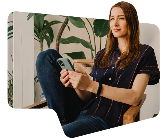 Woman sitting in a wooden chair using a smartphone with plants in the background.